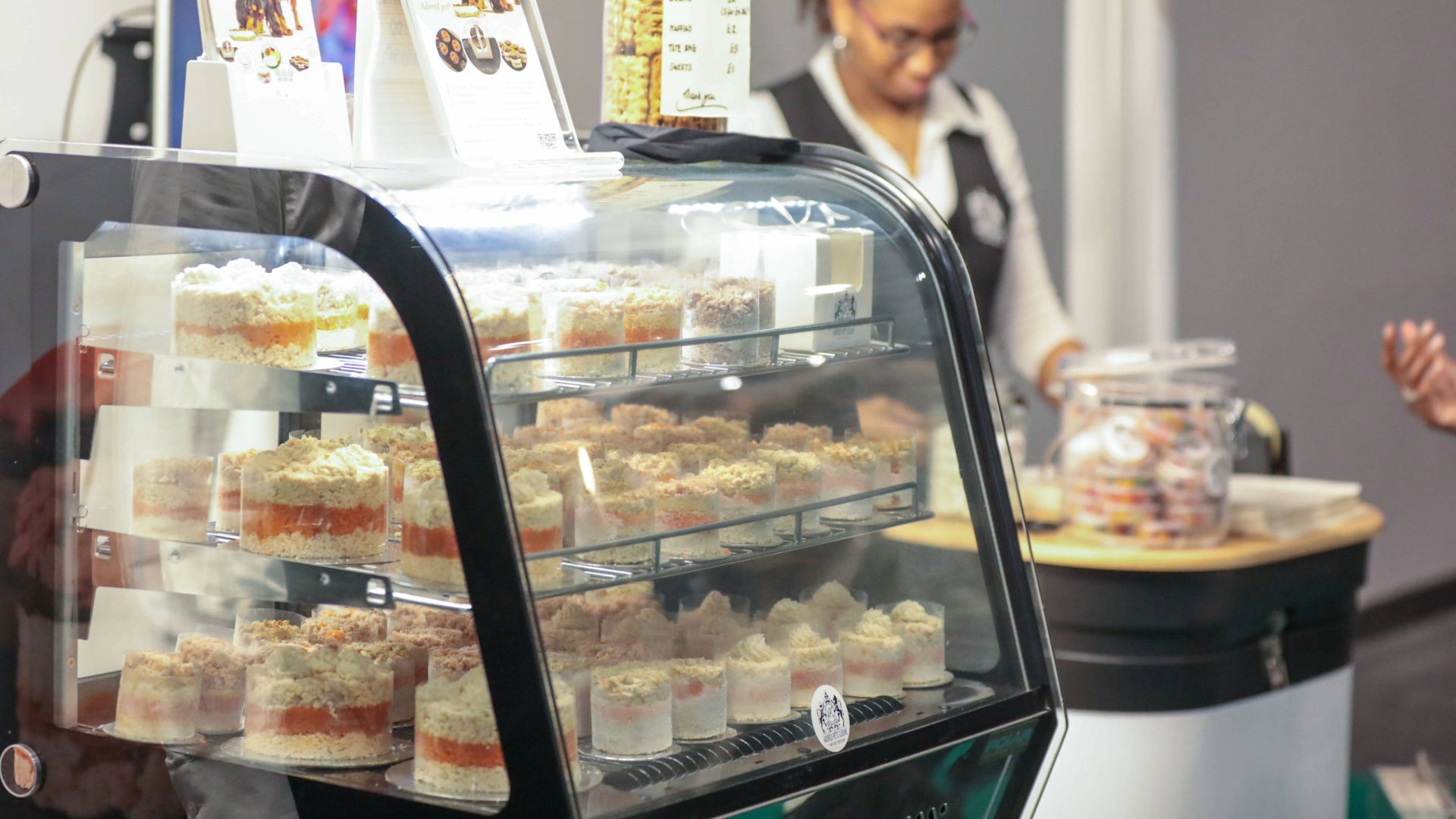 A cabinet full of meat cupcakes for pets of various sizes, with an employee dressed in the background serving a customer at a trade fair stand