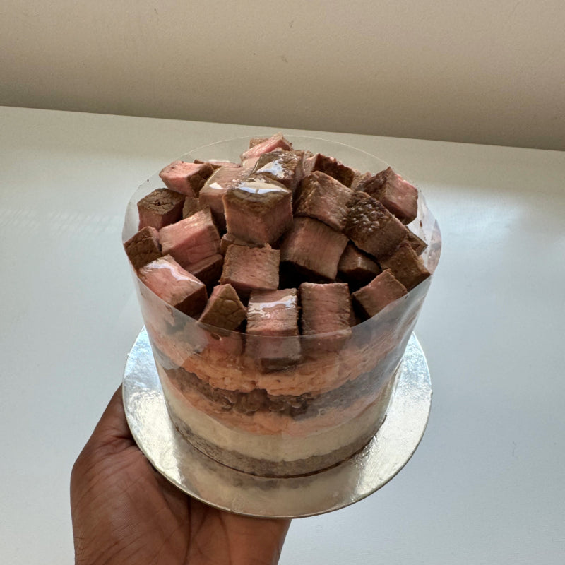 A person holding a steak cake for pets, containing layers of meat and topped with pieces of steak